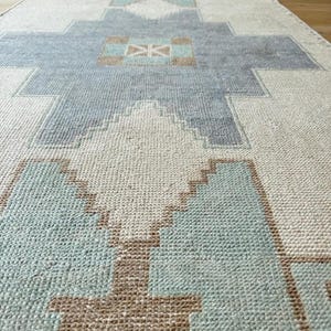 May include: A long, rectangular rug with a geometric pattern in shades of light blue, beige, and brown. The rug features a repeating design of stylized shapes, creating a textured surface. The rug is made of a woven material.