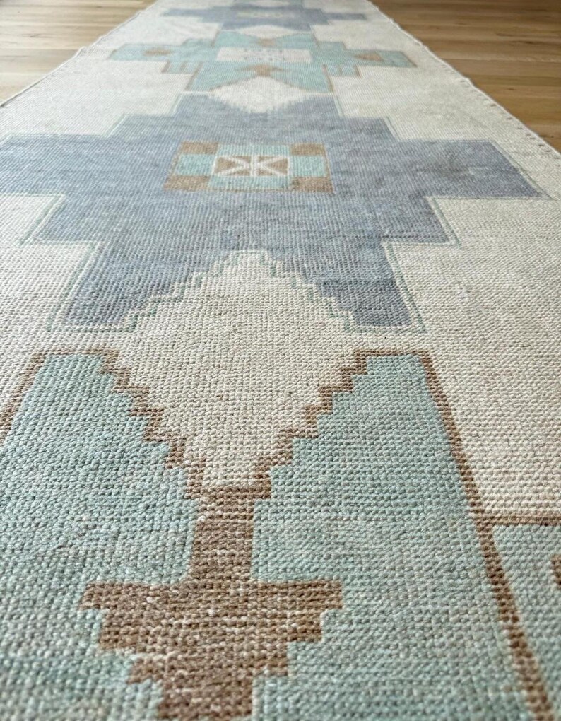 May include: A long, rectangular rug with a geometric pattern in shades of light blue, beige, and brown. The rug features a repeating design of stylized shapes, creating a textured surface. The rug is made of a woven material.