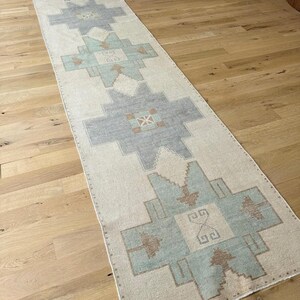 May include: A long, rectangular rug with a cream background and geometric patterns in grey, blue, and brown. The rug is placed on a wooden floor and has a traditional design.