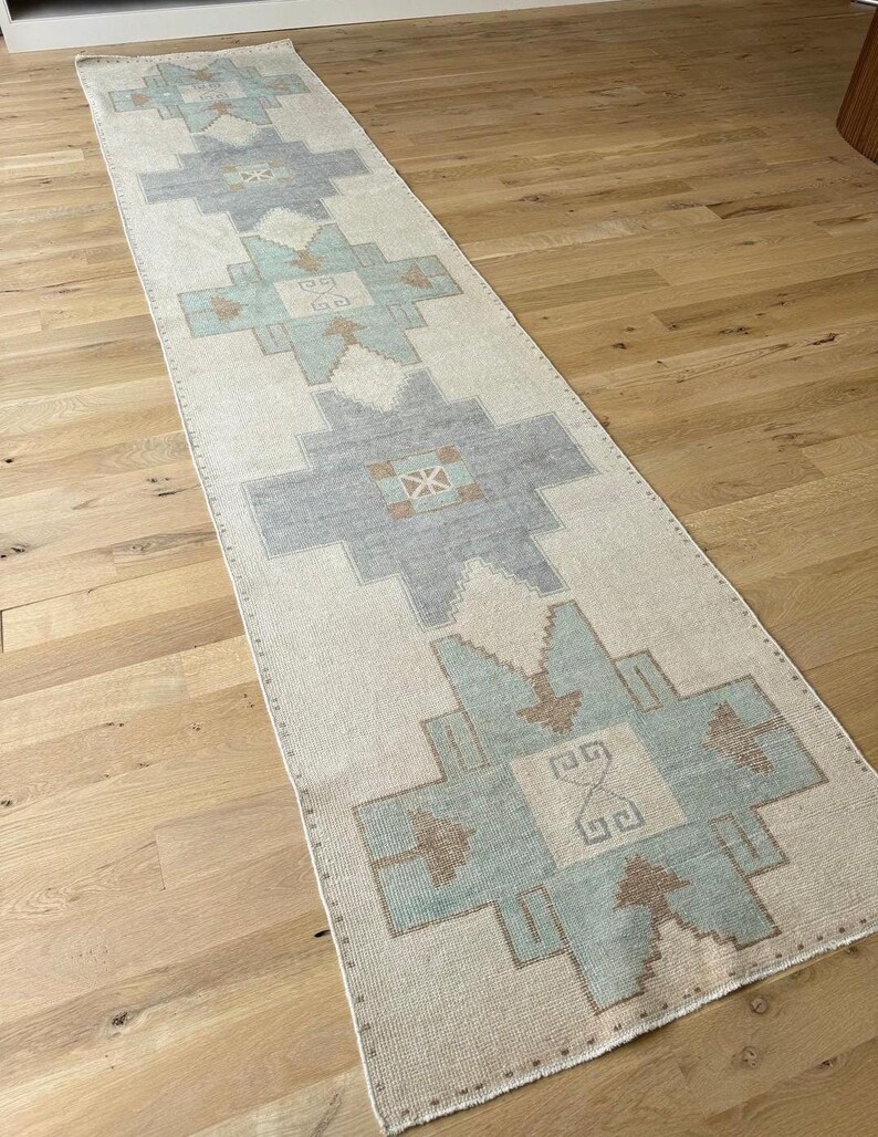 May include: A long, rectangular rug with a cream background and geometric patterns in grey, blue, and brown. The rug is placed on a wooden floor and has a traditional design.