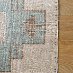 May include: Close-up of a rectangular rug with a geometric pattern. The rug features a light beige background with a repeating design in light blue and brown. The edges are finished with a stitched border. The rug is likely made of woven material.