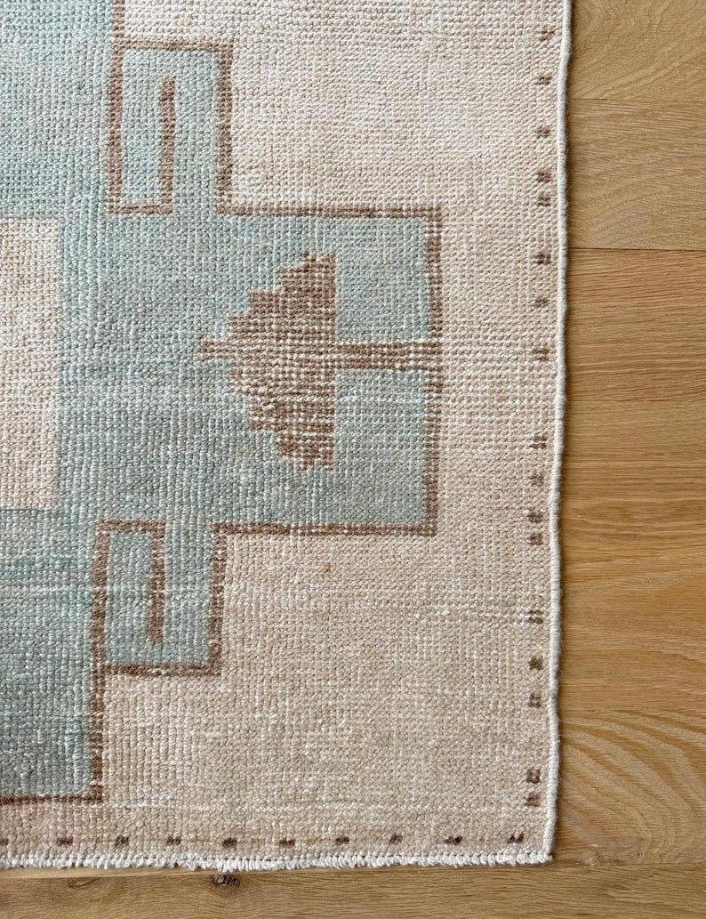 May include: Close-up of a rectangular rug with a geometric pattern. The rug features a light beige background with a repeating design in light blue and brown. The edges are finished with a stitched border. The rug is likely made of woven material.
