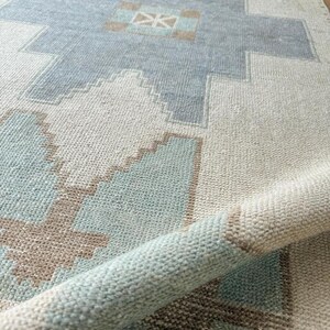 May include: A rolled-up, rectangular rug with a geometric pattern. The rug features a light blue, beige, and brown design. The pattern includes diamond and cross shapes. The rug is made of a woven material.