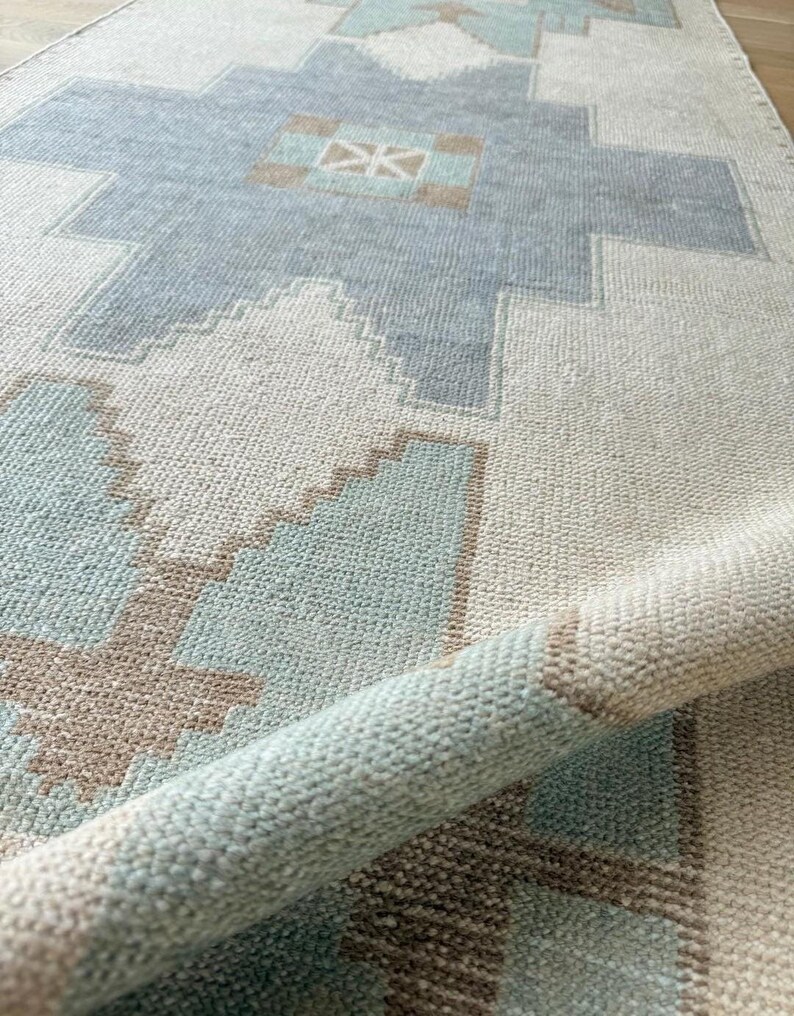 May include: A rolled-up, rectangular rug with a geometric pattern. The rug features a light blue, beige, and brown design. The pattern includes diamond and cross shapes. The rug is made of a woven material.