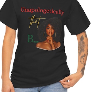 Op de afbeelding: Zwart T-shirt met de tekst "Unapologetically that B..." in rood, geel en groen. Het shirt heeft een illustratie van een vrouw met krullend haar, die een vinger voor haar lippen houdt. Het model draagt een blauwe spijkerbroek.