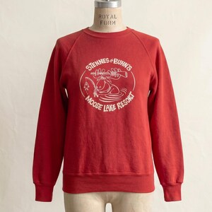 Op de afbeelding: Een rode crewneck sweatshirt met witte tekst en een afbeelding van een eland en een meer. De tekst luidt "Stennes & Buhn's Moose Lake Resort" in een cirkelvormig ontwerp. De sweatshirt wordt op een mannequin getoond.
