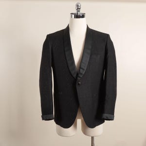 May include: A black blazer with a satin lapel and cuffs, displayed on a mannequin. The blazer has a textured pattern and a single-button closure. The sleeves are cuffed, and the overall style is formal.
