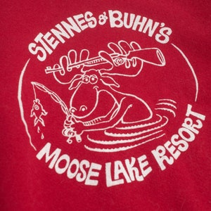 Op de afbeelding: Rood T-shirt met een witte afbeelding van een eland die vist met een geweer en de tekst "Stennes & Buhn's Moose Lake Resort" in een cirkel.