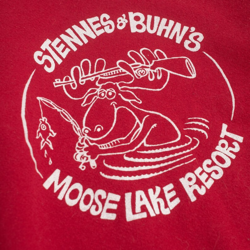 Op de afbeelding: Rood T-shirt met een witte afbeelding van een eland die vist met een geweer en de tekst "Stennes & Buhn's Moose Lake Resort" in een cirkel.