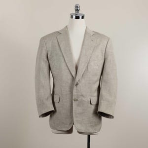 May include: A light beige blazer with a textured weave, featuring a notched lapel, two front buttons, and two flap pockets. The garment is displayed on a mannequin, showcasing long sleeves and a structured fit with a classic tailored design.