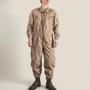 May include: A tan-coloured jumpsuit with a collared neckline, long sleeves, and a full-length zip. It has multiple pockets and a relaxed fit. The model is wearing brown lace-up boots. A practical and stylish garment.