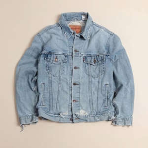May include: Light blue denim jacket with a classic design. Features two chest pockets, two side pockets, and a button-up front. The jacket has a distressed look with frayed edges and a red tag on the chest pocket. A timeless piece for casual wear.