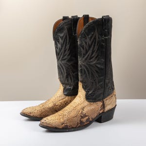 May include: A pair of tall cowboy boots featuring a textured, tan and brown snakeskin-patterned leather foot and a solid black leather shaft. The black shaft is decorated with intricate, tonal stitched wing-like patterns and pull tabs.