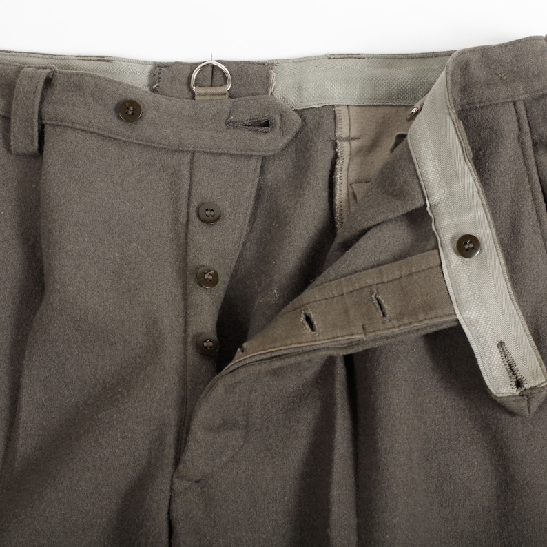 May include: A pair of taupe-colored wool trousers featuring a button-fly closure with four dark brown buttons, a metal D-ring attached to the waistband, belt loops, and a light gray interior lining along the waist and the button placket.
