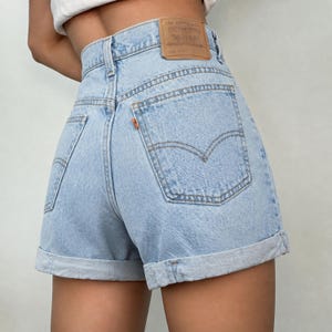 May include: Light blue denim shorts with a high-waisted fit, featuring a tan leather patch on the back waistband with the Levi Strauss & Co. logo, an orange tab on the back pocket, and rolled cuffs at the bottom hem of each leg.