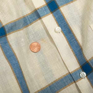 May include: A cream-colored shirt featuring a blue and tan plaid pattern. The garment includes a vertical button placket with two visible white buttons. A copper-colored penny is placed on the fabric to provide a sense of scale for the pattern.