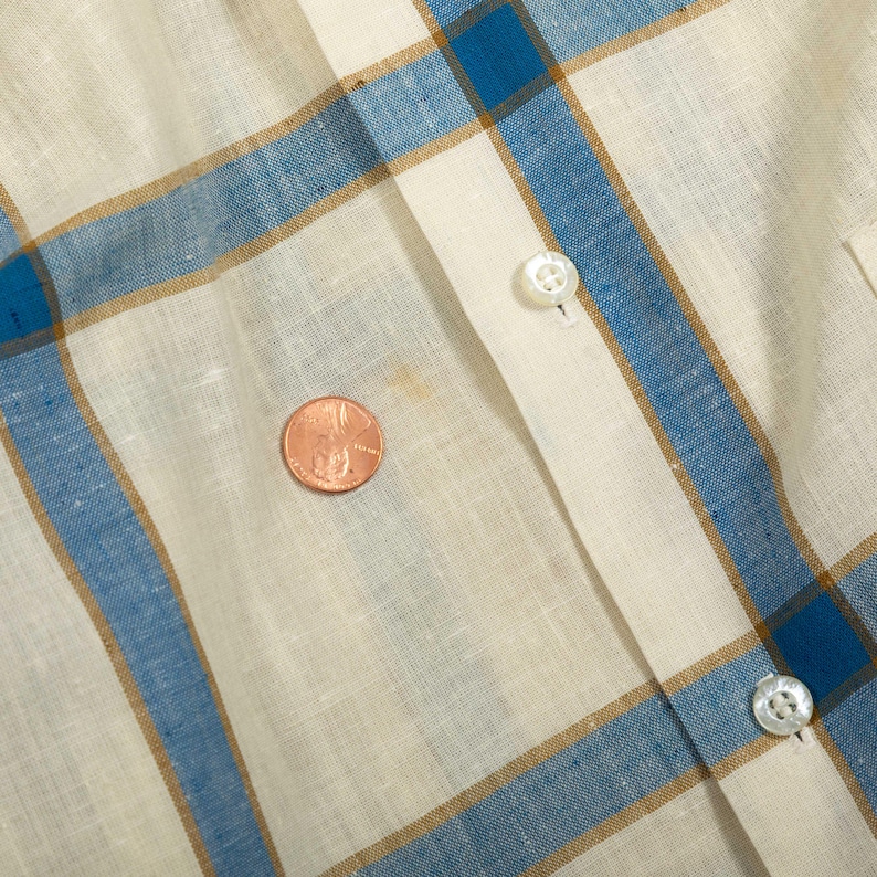 May include: A cream-colored shirt featuring a blue and tan plaid pattern. The garment includes a vertical button placket with two visible white buttons. A copper-colored penny is placed on the fabric to provide a sense of scale for the pattern.