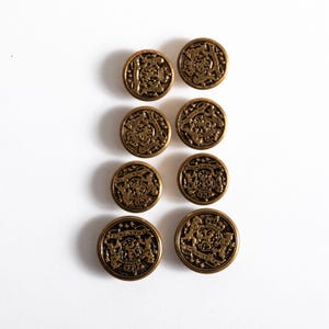 May include: Eight antique brass buttons with a detailed embossed design. The buttons are round and feature a crest-like emblem with intricate patterns and text. The buttons are arranged in a grid pattern against a white background.