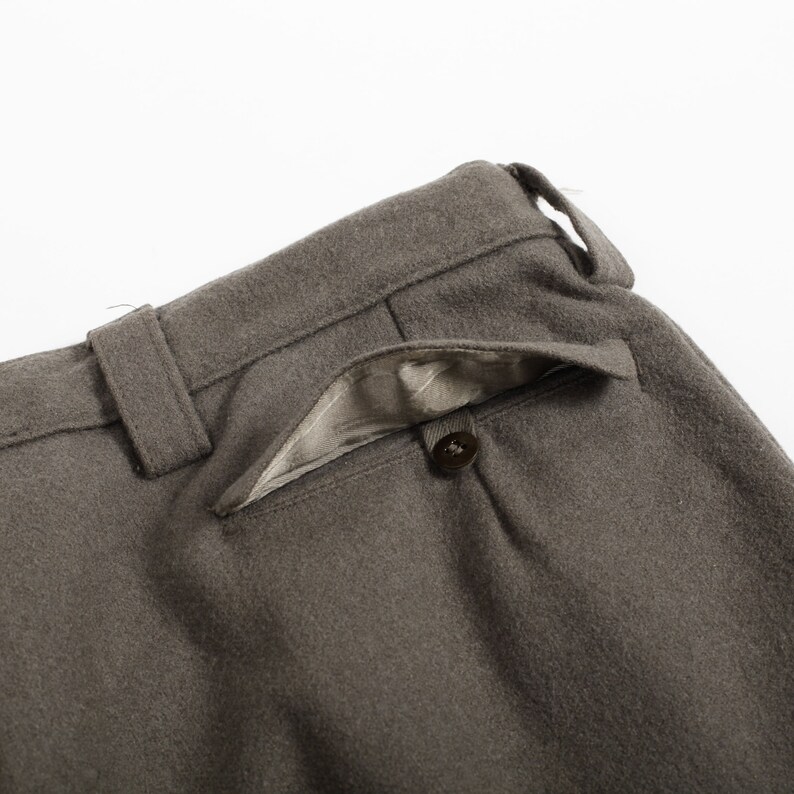 May include: A pair of taupe trousers featuring a textured wool-like fabric, a waistband with belt loops, and a single rear welt pocket secured with a small dark brown button and lined with a light taupe fabric visible at the pocket opening.