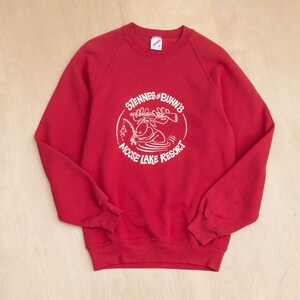 Op de afbeelding: Een rode sweatshirt met een witte afbeelding van een eland die aan het vissen is, met de tekst "Stennes & Buhn's Moose Lake Resort" in een cirkelvormig ontwerp. De sweatshirt heeft een ronde hals en lange mouwen.