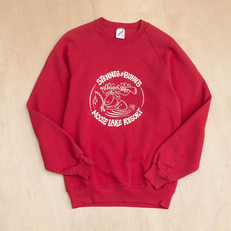 Op de afbeelding: Een rode sweatshirt met een witte afbeelding van een eland die aan het vissen is, met de tekst "Stennes & Buhn's Moose Lake Resort" in een cirkelvormig ontwerp. De sweatshirt heeft een ronde hals en lange mouwen.