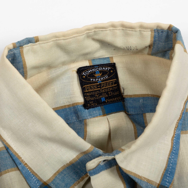 May include: A button-down shirt featuring a cream, blue, and tan plaid pattern. The interior collar displays a black woven label with the text Towncraft Tapered, Penn-Prest, means you NEVER, When Tumble Dried, M, 15-15 1/2, and Penneys.