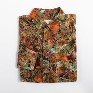 May include: A long-sleeve button-down shirt featuring an abstract, multi-colored print in shades of orange, olive green, brown, and burgundy. The shirt has a pointed collar, a front button placket with dark buttons, and a single chest pocket.