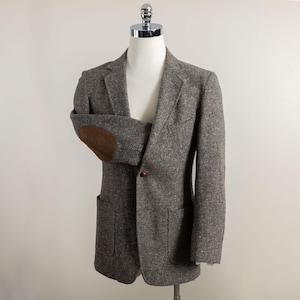 May include: A gray tweed blazer with brown elbow patches and a single brown button closure. The blazer has two front pockets and is displayed on a mannequin. The fabric has a textured appearance.