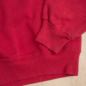 Op de afbeelding: Close-up van een rode sweatshirt. De afbeelding benadrukt de geribbelde manchetten en zoom van het kledingstuk. De stof lijkt een zacht, gebreid materiaal te zijn. De mouwboord is omgeslagen.