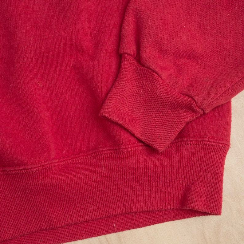 Op de afbeelding: Close-up van een rode sweatshirt. De afbeelding benadrukt de geribbelde manchetten en zoom van het kledingstuk. De stof lijkt een zacht, gebreid materiaal te zijn. De mouwboord is omgeslagen.