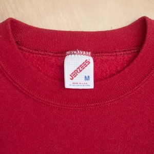 Op de afbeelding: Close-up van een rode sweatshirt met een ronde hals. Het label leest "JERZEES" en "M" voor medium. Het label zegt ook "MADE IN U.S.A." en bevat onderhoudsinstructies. De sweatshirt is gemaakt van een zacht materiaal.