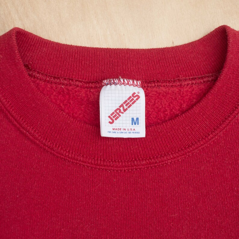Op de afbeelding: Close-up van een rode sweatshirt met een ronde hals. Het label leest "JERZEES" en "M" voor medium. Het label zegt ook "MADE IN U.S.A." en bevat onderhoudsinstructies. De sweatshirt is gemaakt van een zacht materiaal.