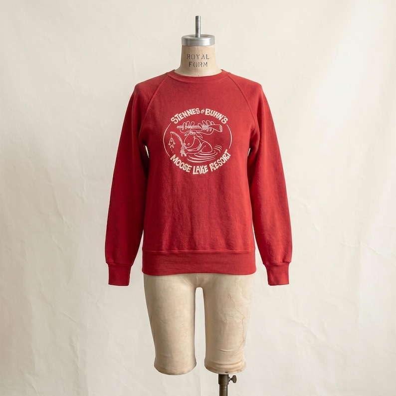 Op de afbeelding: Een rode crewneck sweatshirt met witte tekst en een afbeelding van een eland en een meer. De tekst luidt "Stennes & Buhn's Moose Lake Resort" in een cirkelvormig ontwerp. De sweatshirt wordt op een mannequin getoond.