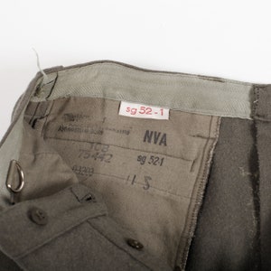 May include: Olive drab trousers featuring an interior waistband with a white tag labeled sg 52-1. The inner lining displays black printed text including NVA, 75442, 03203, and sg 52-1. The garment includes a metal D-ring attachment and brown buttons.