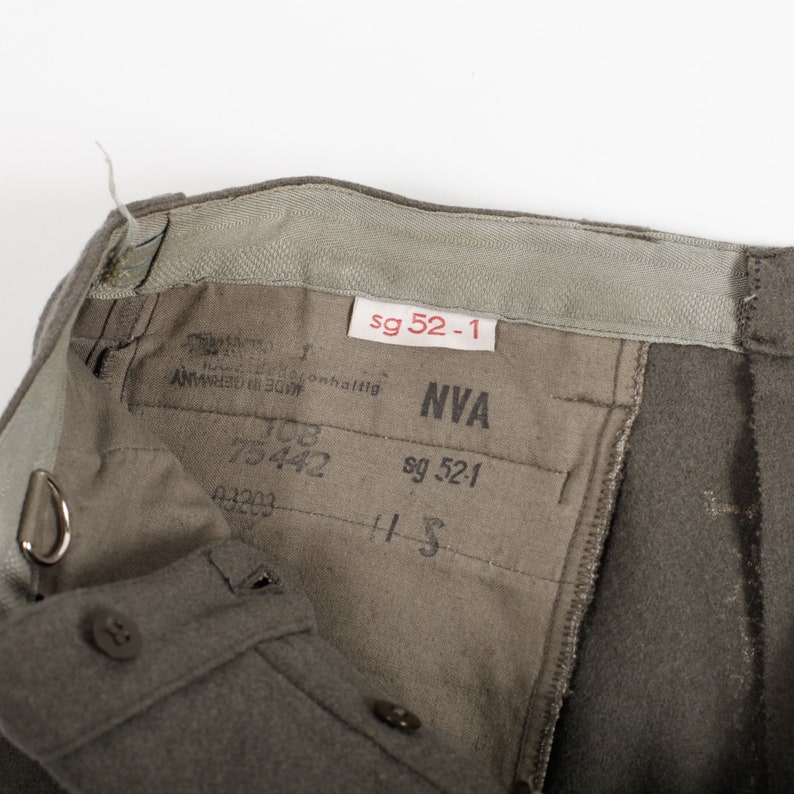 May include: Olive drab trousers featuring an interior waistband with a white tag labeled sg 52-1. The inner lining displays black printed text including NVA, 75442, 03203, and sg 52-1. The garment includes a metal D-ring attachment and brown buttons.