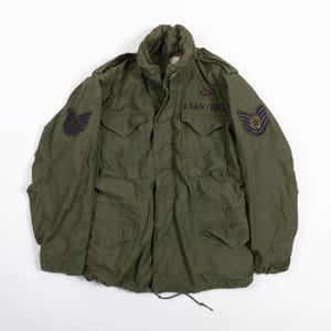 以下が含まれることがあります： ハイカラー、ボタン付きフラップのフロントポケット4つ、長袖を備えたオリーブグリーンのフィールドジャケット。胸元にはダークブルーで「U.S. AIR FORCE」の文字が記され、両袖にはブルーとゴールドのミリタリーワッペンがあしらわれています。
