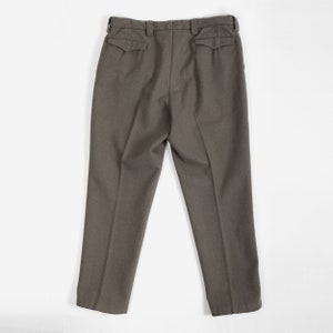 May include: A pair of olive drab trousers featuring a high-waisted design with belt loops, two rear pockets with pointed flaps, and a straight-leg cut. The pants are constructed from a textured fabric and display a clean, structured appearance.