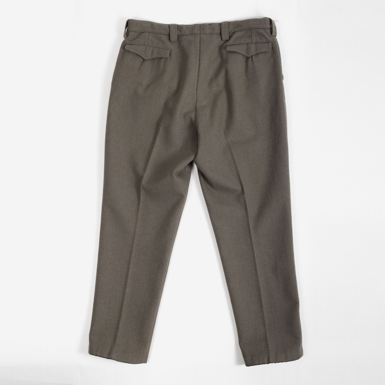 May include: A pair of olive drab trousers featuring a high-waisted design with belt loops, two rear pockets with pointed flaps, and a straight-leg cut. The pants are constructed from a textured fabric and display a clean, structured appearance.