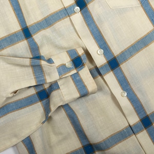 May include: A long-sleeved button-down shirt featuring a plaid pattern with intersecting blue and tan lines on an off-white background. The shirt includes a front placket with white buttons and a matching buttoned cuff on the sleeve.