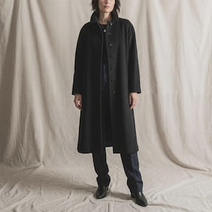 May include: A long, black coat with a button-up front and a high collar. The coat is worn over dark pants and black shoes. The overall style is minimalist and classic, suitable for cooler weather.