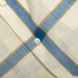 May include: A button-down shirt featuring a woven fabric with a plaid pattern in cream, blue, and tan. The garment includes a vertical placket with white buttons and a chest pocket, showcasing a textured weave throughout the material.