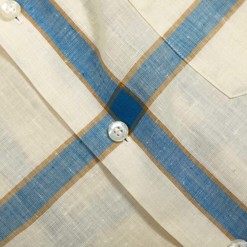 May include: A button-down shirt featuring a woven fabric with a plaid pattern in cream, blue, and tan. The garment includes a vertical placket with white buttons and a chest pocket, showcasing a textured weave throughout the material.