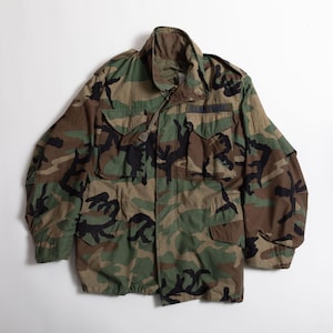 May include: A camouflage field jacket featuring a woodland pattern in shades of green, brown, and black. The jacket includes a high collar, long sleeves, two chest pockets with flaps, and a patch above the right pocket reading U.S. AIR FORCE.