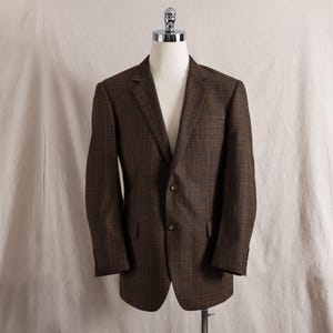 May include: A brown plaid blazer with a two-button closure, displayed on a mannequin. The jacket features a classic notched lapel, two front pockets, and buttoned cuffs. The fabric has a subtle checkered pattern.