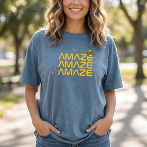 Peut inclure: T-shirt bleu avec le mot "AMAZE" en jaune, répété trois fois, avec un motif de planète et d'étoiles. Le t-shirt est de couleur unie et a des manches courtes.