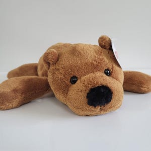 May include: A brown plush teddy bear with a black nose and black eyes. The bear is lying down with its arms and legs extended. A small tag is attached to the bear.