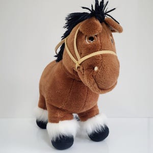May include: A brown plush horse with a black mane and tail, and white fur on its legs. The horse has a tan bridle and large, expressive eyes. The toy is standing on a white surface.