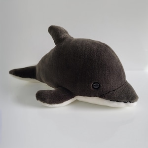 May include: A soft toy dolphin in grey and white. The plushie has a dark grey body, a dorsal fin, and a tail. The underside and edges of the fins are white. The dolphin has a small, black eye.