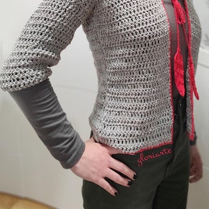 Puede incluir: Un cárdigan gris de punto de ganchillo con ribete rojo y una corbata roja en la parte delantera. El cárdigan tiene un corte holgado y está hecho con un punto texturizado.