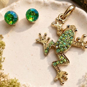 May include: A gold-toned metal frog pendant featuring a back covered in small, sparkling green crystals, accompanied by a pair of matching round stud earrings, each set with a single faceted, iridescent green crystal stone.